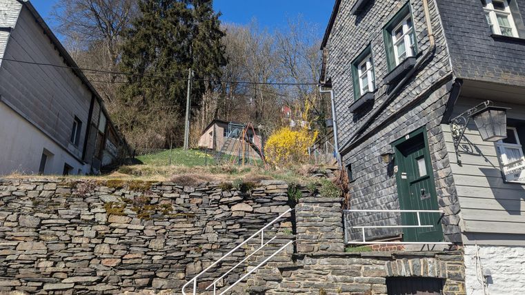 Ein steiler Weg mit einer Steinmauer und zwei Häusern. Im Hintergrund sind Bäume und eine Wiese mit gelben Blumen zu sehen.