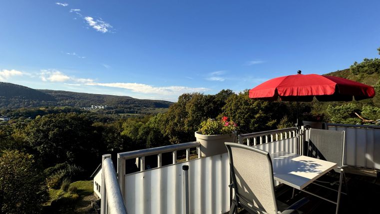 Ein Balkon mit einem Tisch und Stühlen bietet einen schönen Ausblick auf die umliegenden Berge und das Tal. Eine rote Sonnenschirm steht bereit für Schatten.