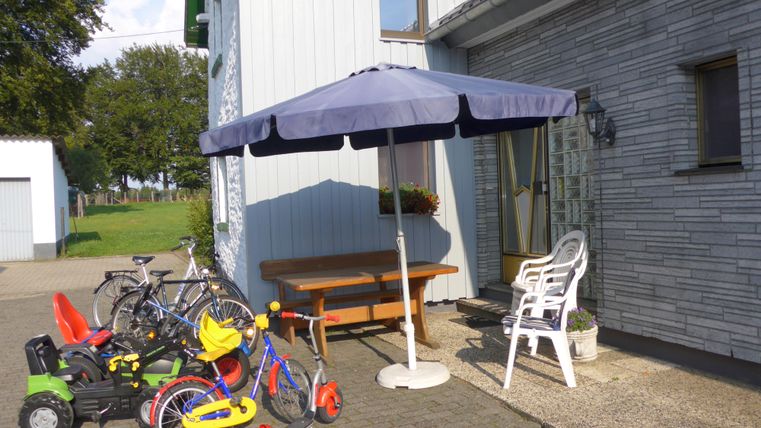 Ein Eingang mit einem Sonnenschirm und einem Tisch. Neben dem Eingang stehen verschiedene Spielzeuge und Fahrräder.