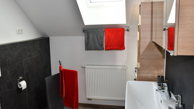 Ein modernes Badezimmer mit einem Fenster und viel Tageslicht. Die Wände sind in neutralen Farben gehalten, ergänzt durch rote und graue Handtücher.