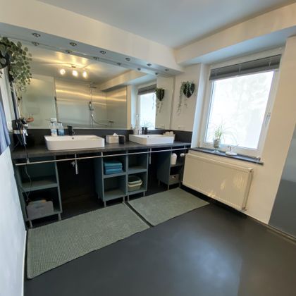 Ein modernes Badezimmer mit zwei Waschtischen und großen Spiegeln. Fenster sorgen für viel Tageslicht und Pflanzen verleihen dem Raum eine frische Atmosphäre.