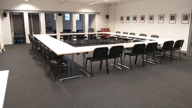 Ein saal mit einem rechteckigen Tisch und schwarzen Stühlen. Die Wände sind mit Bildern dekoriert, und es gibt große Fenster, die natürliches Licht hereinlassen.
