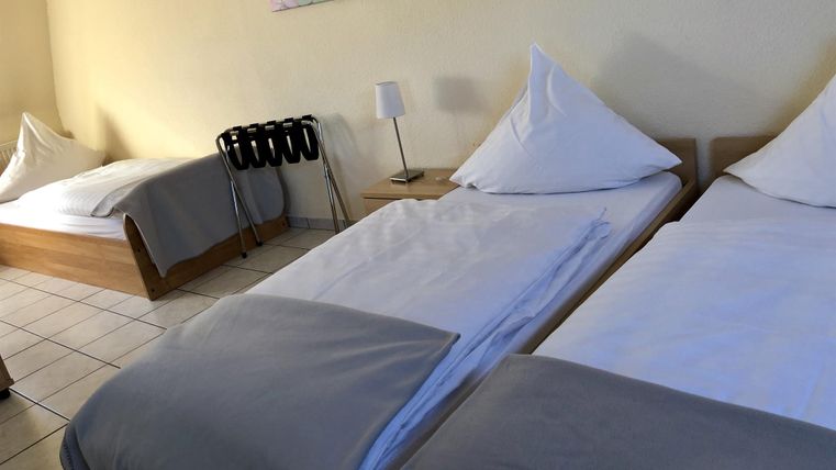 Ein einfaches Zimmer mit drei Betten und neutraler Einrichtung. Die Wände sind hell, und es gibt ein Gemälde als Dekoration.