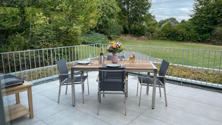 Ein eleganter Essbereich auf einer Terrasse mit einem Tisch und Stühlen. Blick auf einen gepflegten Garten mit Bäumen im Hintergrund.