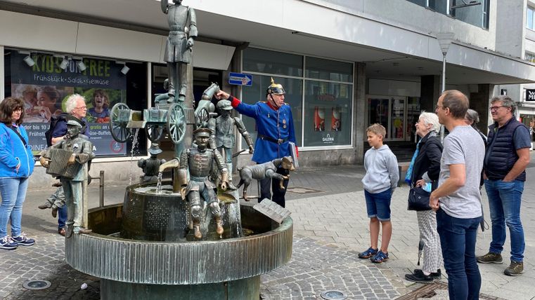 Ein Brunnen mit einer Bronzeskulptur von Figuren, die eine Geschichte erzählen. Eine Gruppe von Menschen steht daneben und schaut zu.