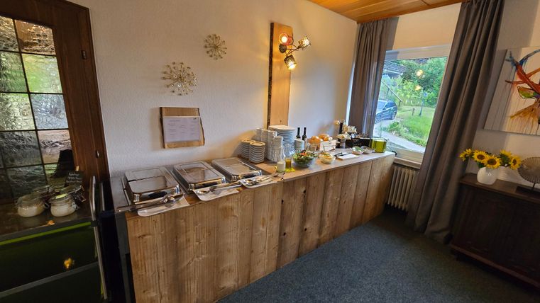 Ein Frühstücksbuffet mit verschiedenen Speisen steht auf einem hölzernen Tisch. Durch ein Fenster ist eine grüne Gartenlandschaft zu sehen.