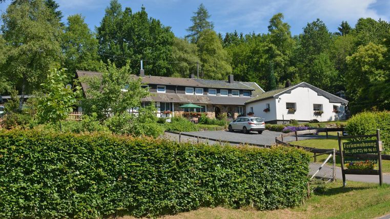 Ein idyllisches Hotel inmitten von Bäumen mit einer begrünten Umgebung. Parkplatz vor dem Gebäude und einladende Terrassenbereiche sind sichtbar.