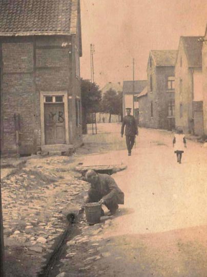 Eine historische Straßenansicht mit Steinen und alten Häusern. Ein Mann arbeitet am Boden, während eine Person und ein Kind vorbeigehen.