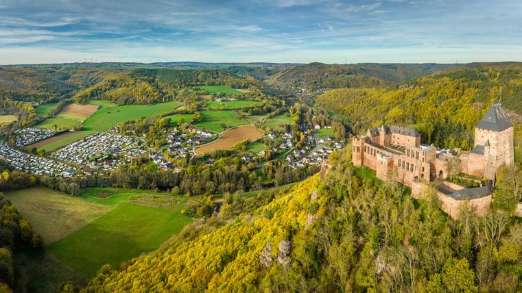 Eine beeindruckende Aussicht auf eine Burg, umgeben von grünen Wäldern und sanften Hügeln. Im Vordergrund ist ein Campingplatz in einem Tal zu sehen.