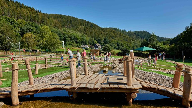 Ein malerischer Holzwagen führt über einen kleinen Wasserlauf. Im Hintergrund sind grüne Hügel und Besucher zu sehen, die die Natur genießen.