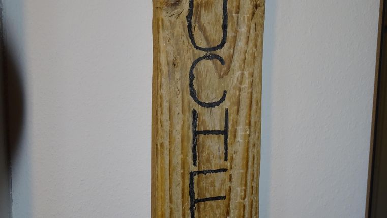 Ein hölzerner Schild mit dem Wort "KÜCHE" in großen Buchstaben. Der Schild ist an einer Wand befestigt.