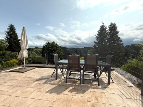 Eine Terrasse mit einem Tisch und Stühlen, umgeben von grünen Bäumen. Der Himmel ist teilweise bewölkt und die Landschaft ist gut sichtbar.