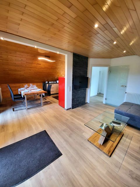 Ein modernes Wohnzimmer mit Holzdecke und einem gemütlichen Sofa. Der Essbereich ist mit einem Tisch und schwarzen Stühlen ausgestattet.