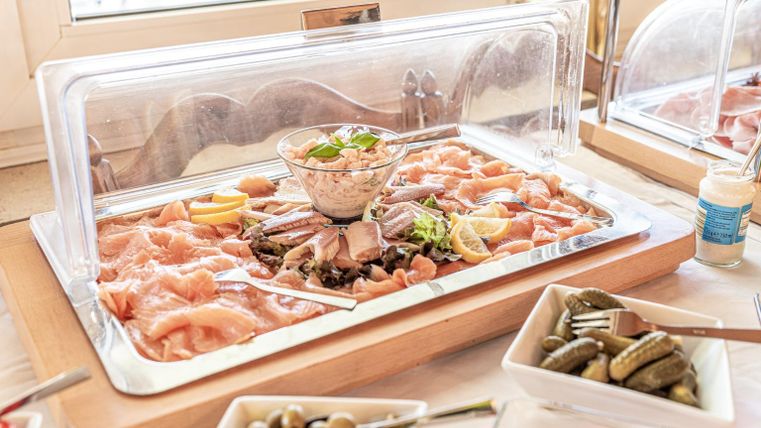 Ein Buffet mit verschiedenen Fischsorten und Garnelen, garniert mit Kräutern und Zitronen. Neben dem Fisch befinden sich Schalen mit Oliven und Gewürzgurken.