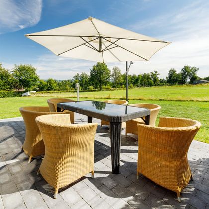 Ein gemütlicher Essbereich im Freien mit einem großen Tisch und sechs Korbstühlen. Ein Sonnenschirm schützt vor der Sonne, umgeben von einer grünen Wiese.