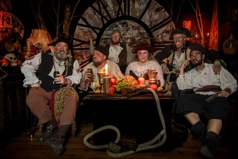 Eine Gruppe von Piraten sitzt um einen Tisch mit Lebensmitteln und Getränken. Die Atmosphäre ist festlich und die Umgebung ist im Piratenstil dekoriert.