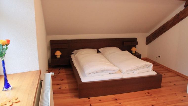 Ein gemütliches Schlafzimmer mit einem Doppelbett und Holzfußboden. Auf dem Nachttisch stehen zwei Lampen und eine Vase mit Blumen.