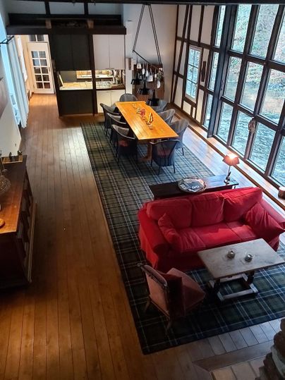 Ein modernes Wohnzimmer mit einem roten Sofa und einem großen Holztisch. Große Fenster lassen viel Licht herein und bieten einen Blick ins Freie.