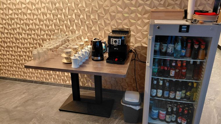 Eine Kaffeestation mit Gläsern und einer Kaffeemaschine steht auf einem Tisch. Nebenan befindet sich ein Kühlschrank mit verschiedenen Getränken.