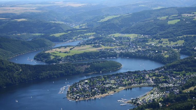 Eine malerische Landschaft mit einem weitläufigen See und umliegenden Hügeln. Das grüne Ufer ist von kleinen Städten und Booten gesäumt.