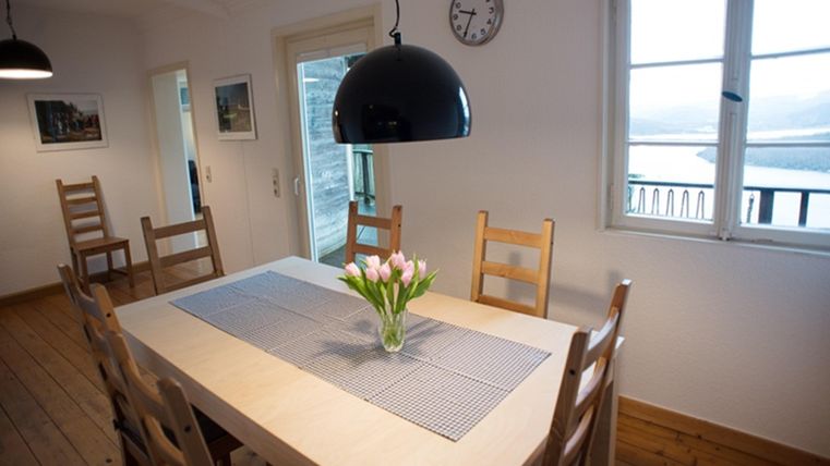 Ein gemütlicher Essbereich mit einem großen Tisch und Holzstühlen. Auf dem Tisch steht eine Vase mit Blumen und der Raum hat große Fenster mit Blick auf die Landschaft.