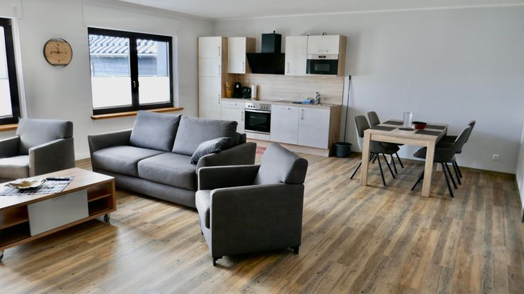 Ein modernes Wohnzimmer mit einem grauen Sofa und einem Holztisch. Die offene Küchenecke ist gut ausgestattet und das Licht fällt durch die großen Fenster.