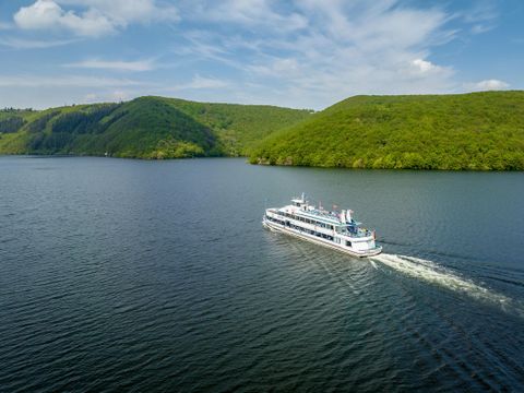 Ein Schiff fährt ruhig über einen See, umgeben von grünen Hügeln. Der Himmel ist blau mit einigen Wolken.